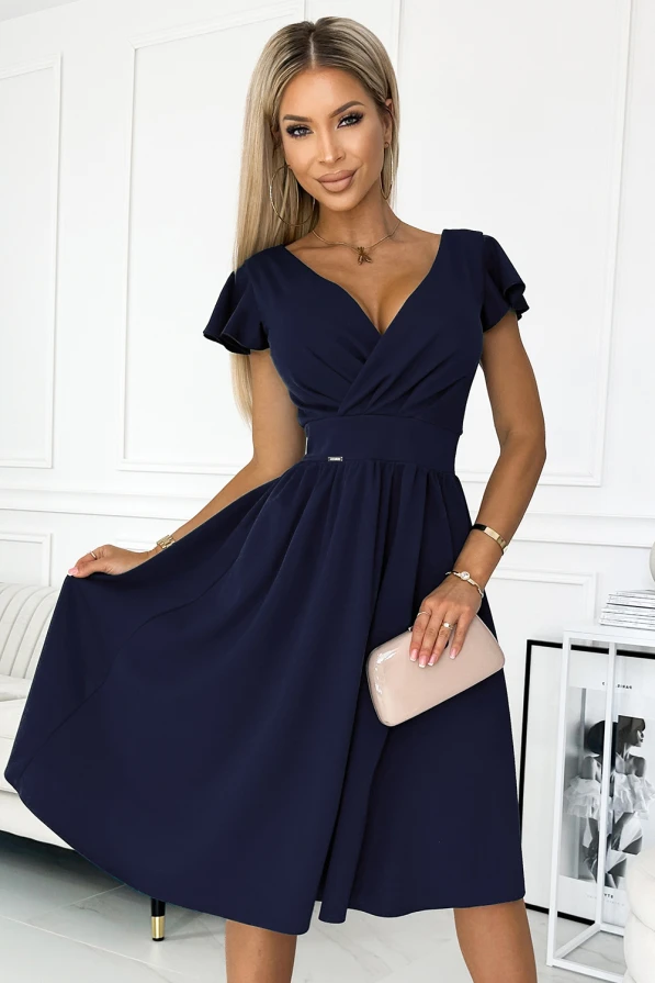 Na obrázku je elegantní tmavě modré šaty s krátkými rukávy a výstřihem do V, které mají jemně řasenou sukni. Modelka drží béžovou kabelku a stojí v moderním interiéru.
