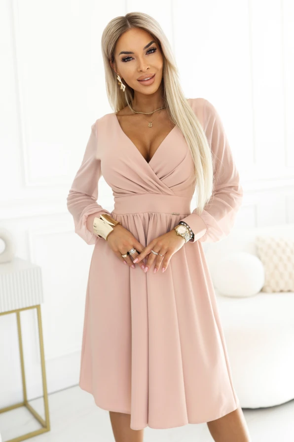 Modelka představuje elegantní béžové midi šaty s kopertovým výstřihem, rozšířenou sukní a lehkými průhlednými rukávky. Šaty jsou ideální na svatbu, komunio, křtiny a rodinné slavnosti, krásně zdůrazňují pas a ženskou siluetu.