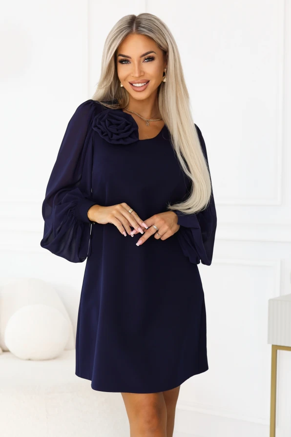 Modelka v tmavě modrých šatech NELIA značky Numoco trapézového střihu těsně nad kolena, s ozdobnou růží u výstřihu a dlouhými průsvitnými rukávy zakončenými širokými manžetami s knoflíkem.
