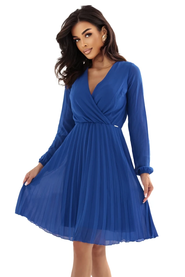 Modelka prezentuje elegancką chabrową sukienkę z kopertowym dekoltem i plisowanym dołem. Stylizacja marki Numoco została sfotografowana w jasnym, nowoczesnym wnętrzu, co podkreśla lekkość i szyk kreacji.