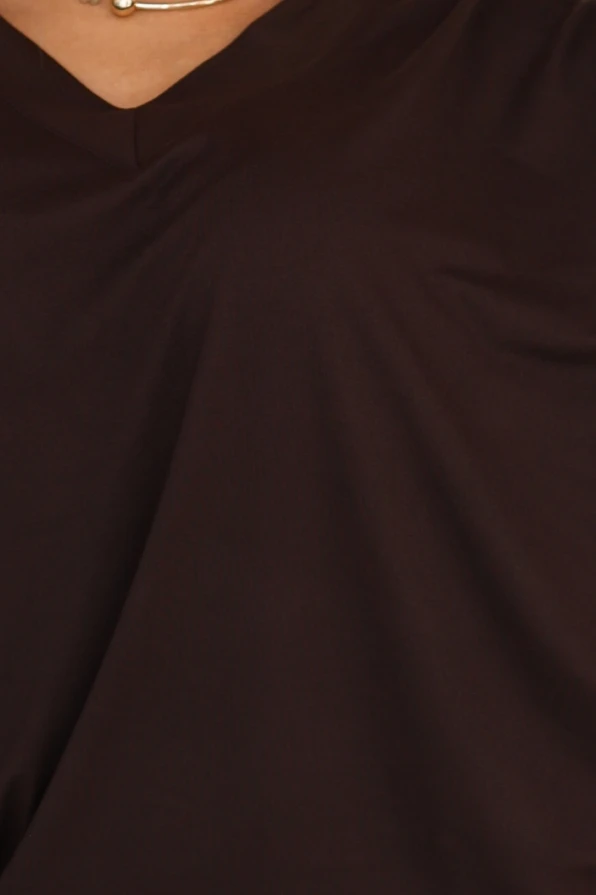 Elegancka kobieta o jasnych włosach w brązowej, zbluzowanej sukience z dekoltem w serek i rękawami typu nietoperz, pozująca w jasnym, minimalistycznym wnętrzu z białymi meblami.