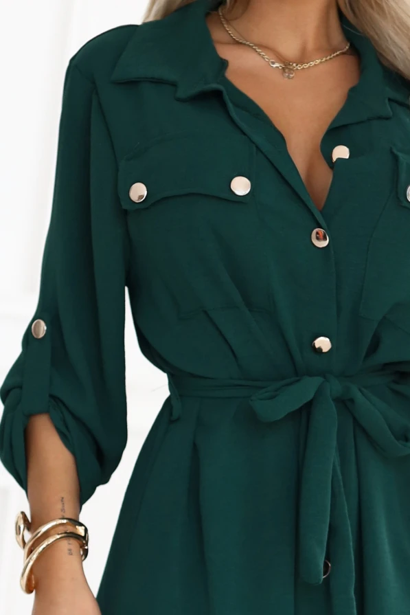 Modelka prezentuje klasyczną szmizjerkę w kolorze zielonym z paskiem i praktycznymi detalami, podkreślającą kobiecą sylwetkę.