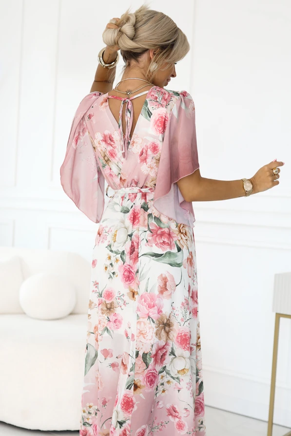 Modelka prezentuje zwiewną sukienkę maxi w romantyczny, kwiatowy wzór w odcieniach różu. Jej subtelna poza podkreśla lekkość tkaniny oraz kobiecy fason z wiązaniem w pasie i luźnymi rękawkami.