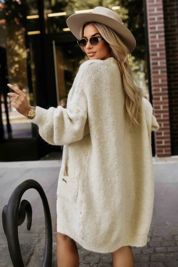 Modelka prezentuje długi kardigan damski z miękkiej dzianiny w kolorze beżowym, z kieszeniami i ozdobną broszką – Numoco Basic.