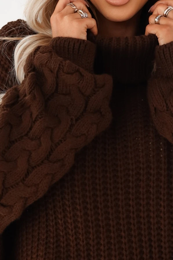 Kobieta w ciemnobrązowym swetrze oversize z golfem i grubym splotem warkoczowym na rękawach, zestawionym z brązową skórzaną spódnicą mini i kapeluszem.