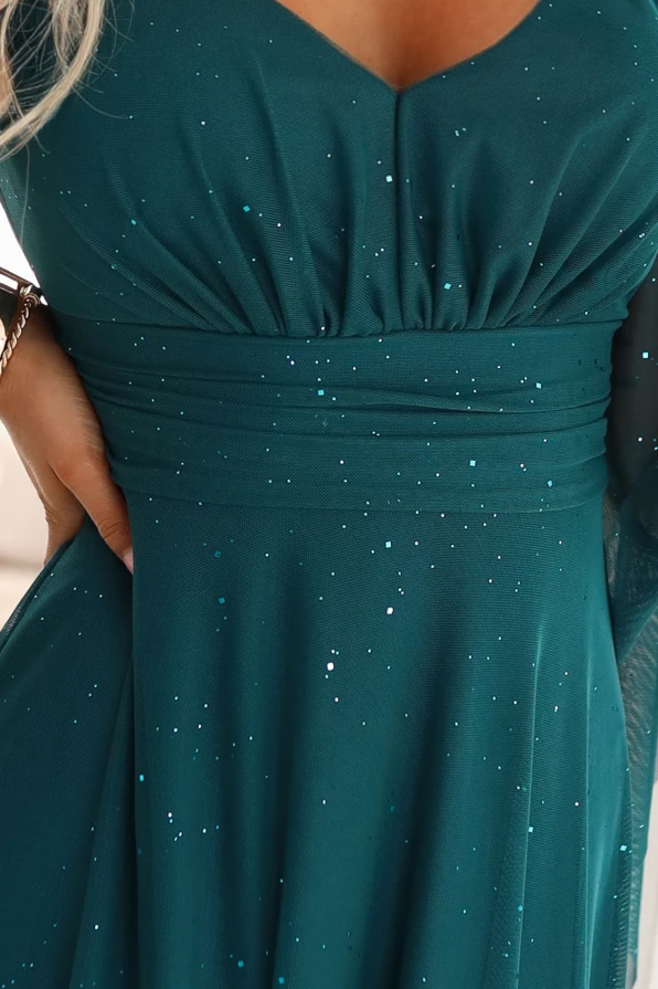 Modelka w szmaragdowej wieczorowej sukience maxi z brokatowym połyskiem i rękawami typu motyl w jasnym wnętrzu.